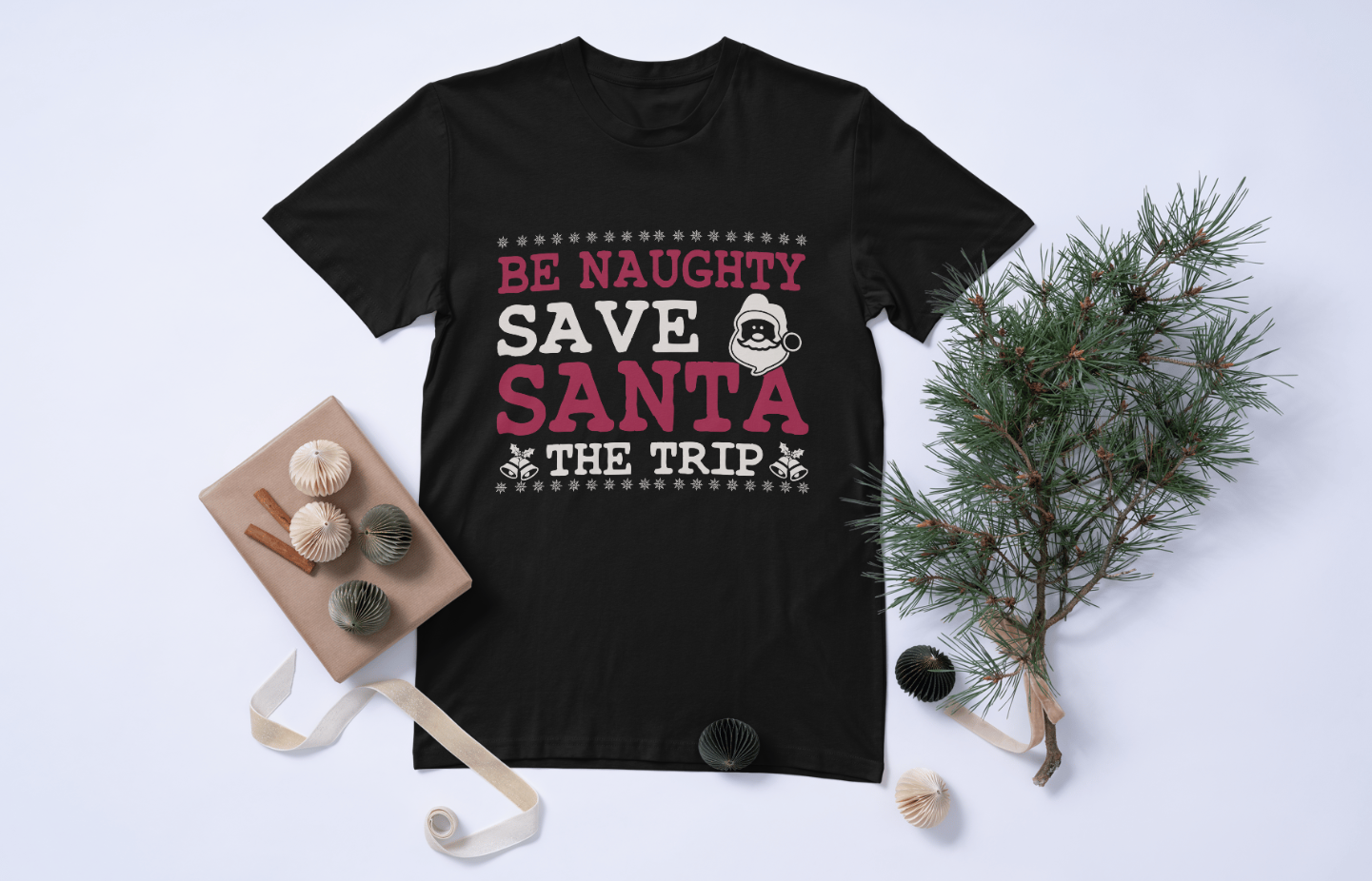 Black short-sleeve t-shirt with a festive graphic design laid out flat on a white surface, accompanied by decorative items and be naughty save santa the trip text.