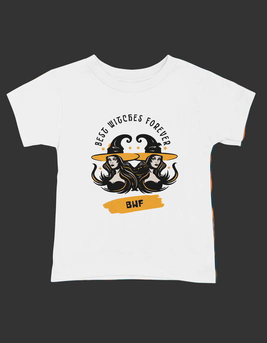 A white short sleeve t-shirt with a graphic print that reads 'Best Witches Forever' and includes images of two witch hats and a cauldron, surrounded by a fiery background.